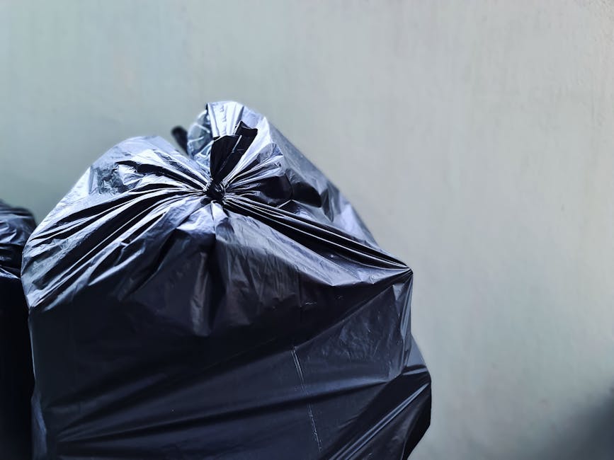 A close-up view of several black plastic rubbish bags with tightly gathered tops, placed against a plain light grey wall. The bags are made of glossy, crinkled plastic material, and they are stacked o