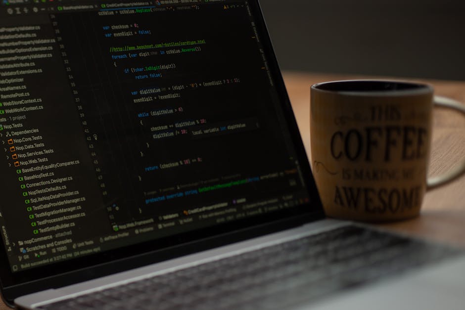 A man with short brown hair, glasses, and a beard is seated at a wooden desk in front of two large computer monitors. The monitors display lines of programming code with syntax highlighting, featuring
